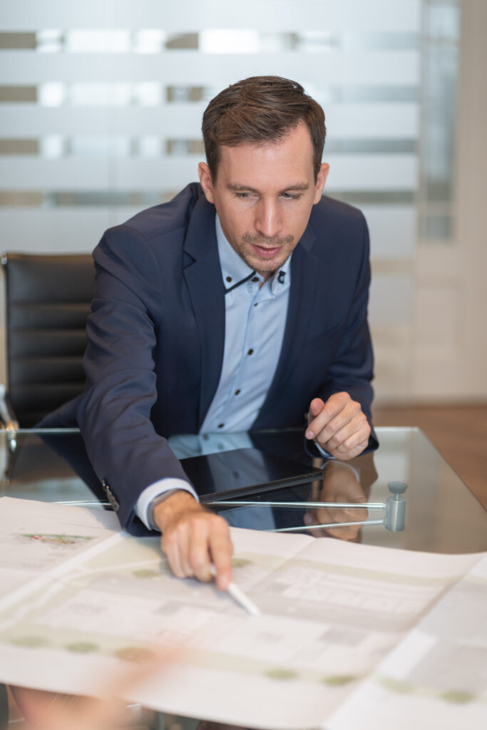 Geschäftsführer der SDH-Immobilien GmbH & Co. KG erläutert Baupläne im Meeting Geschäftsführer der SDH-Immobilien GmbH & Co. KG sitzt im Besprechungsraum und erklärt mit einem Stift Baupläne auf dem Tisch.