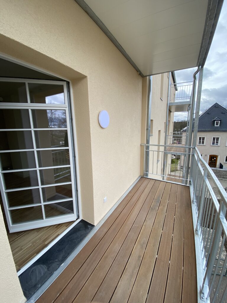Überdachter Balkon mit Holzdielen und Metallgeländer an einem Neubau in der Otto-Schimmel-Straße 11, mit Zugang über große Fenstertür, angeboten von der SDH-Immobilien GmbH & Co. KG.