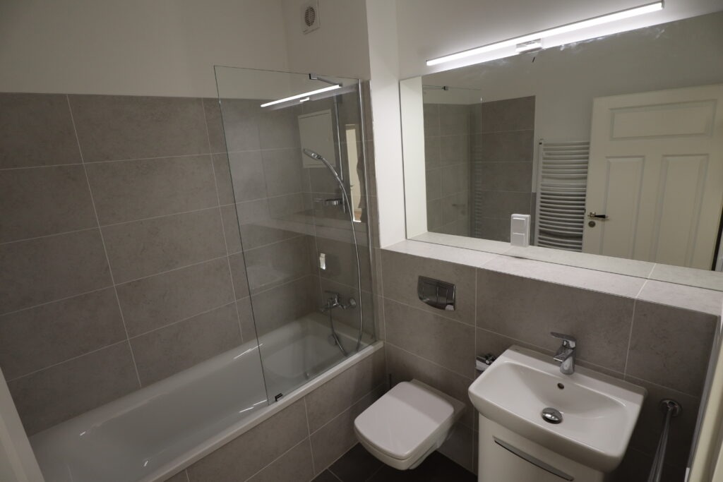 Modernes Badezimmer mit Badewanne und Glasduschabtrennung, grauen Fliesen, Wand-WC, Waschbecken und großem Spiegel in einer Wohnung an der Gießerstraße, angeboten von der SDH-Immobilien GmbH & Co. KG.