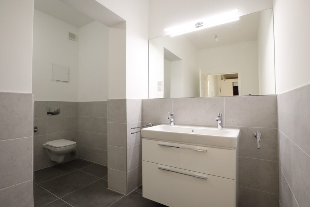 Modernes Badezimmer mit Badewanne und Glasduschabtrennung, grauen Fliesen, Wand-WC, Waschbecken und großem Spiegel in einer Wohnung an der Gießerstraße, angeboten von der SDH-Immobilien GmbH & Co. KG.