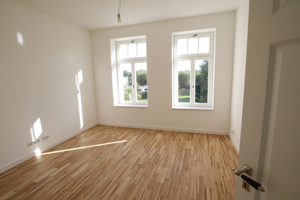 Helles Zimmer mit zwei hohen Fenstern, Blick ins Grüne und neu verlegtem Parkettboden in einer Wohnung in der Heinrich-Lorenz-Straße 11, angeboten von der SDH-Immobilien GmbH & Co. KG.